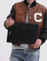 Person wearing a brown and black varsity jacket with a large 'C' patch, holding a black bag, paired with distressed jeans